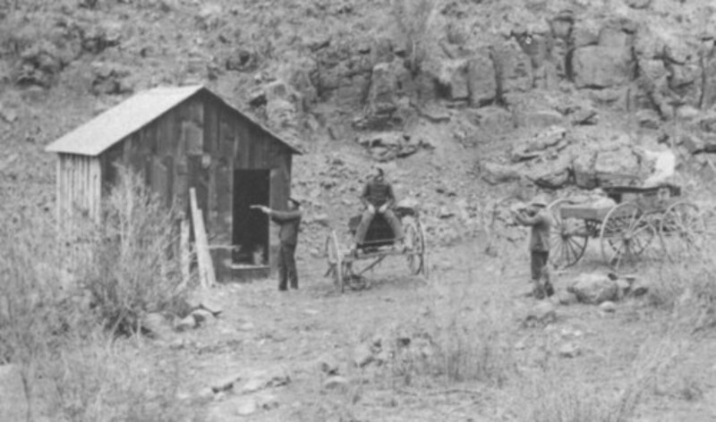 two standing men pointing guns at small cabin while man and woman sit in two buckgoards